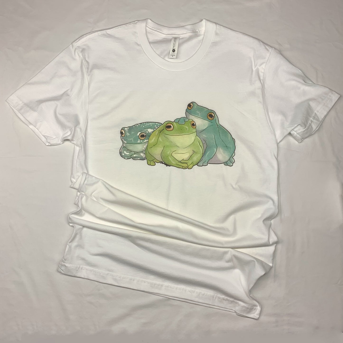 A white, short-sleeve t-shirt with an illustration of three white's tree frogs, each a different color variation, sitting side by side