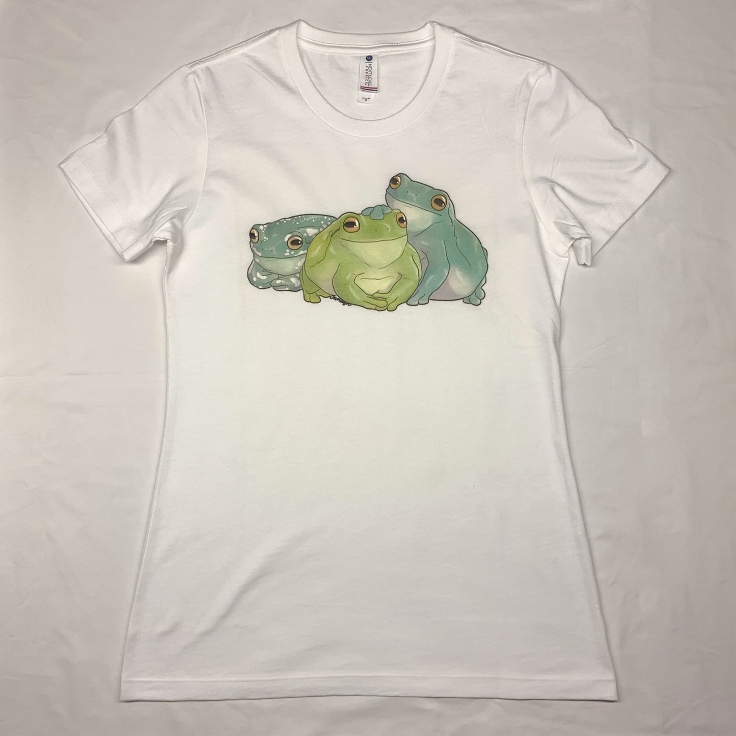 A white, short-sleeve t-shirt with an illustration of three white's tree frogs, each a different color variation, sitting side by side