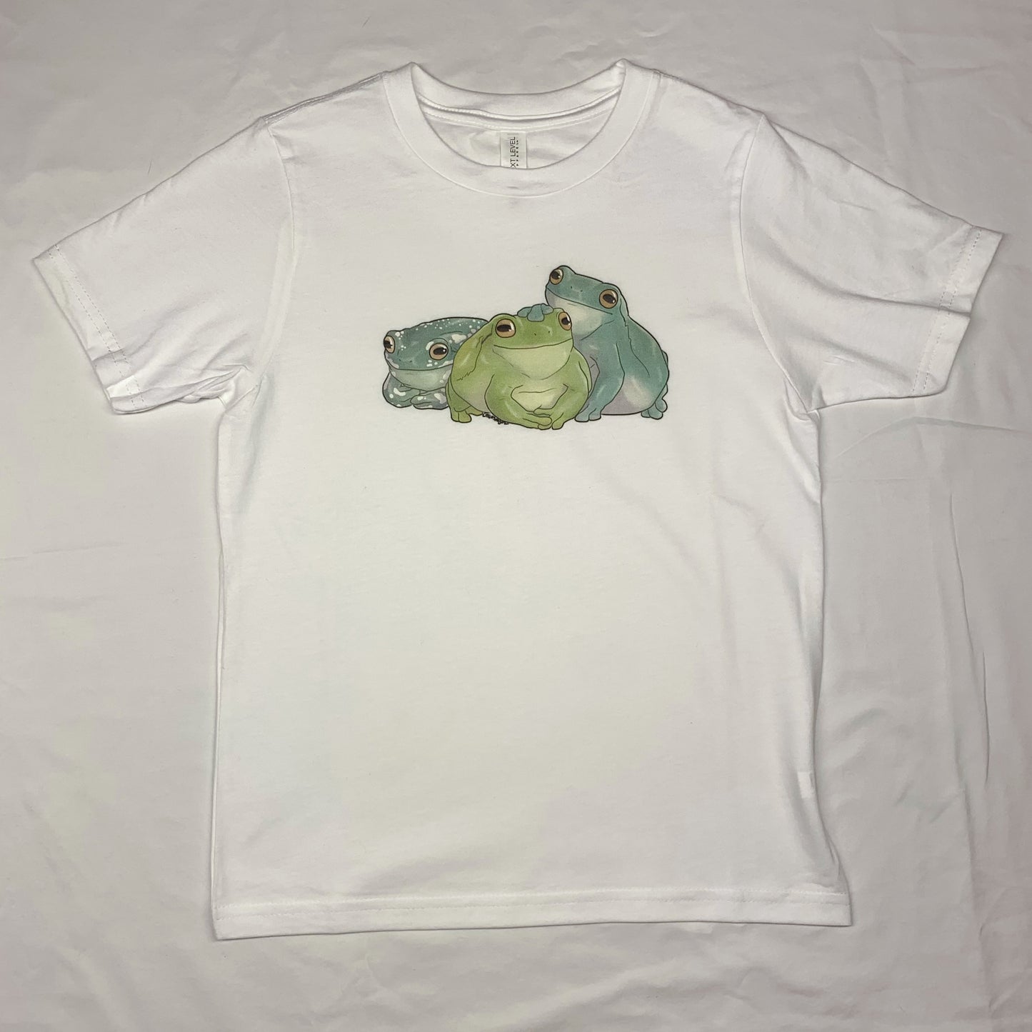 A white, short-sleeve t-shirt with an illustration of three white's tree frogs, each a different color variation, sitting side by side
